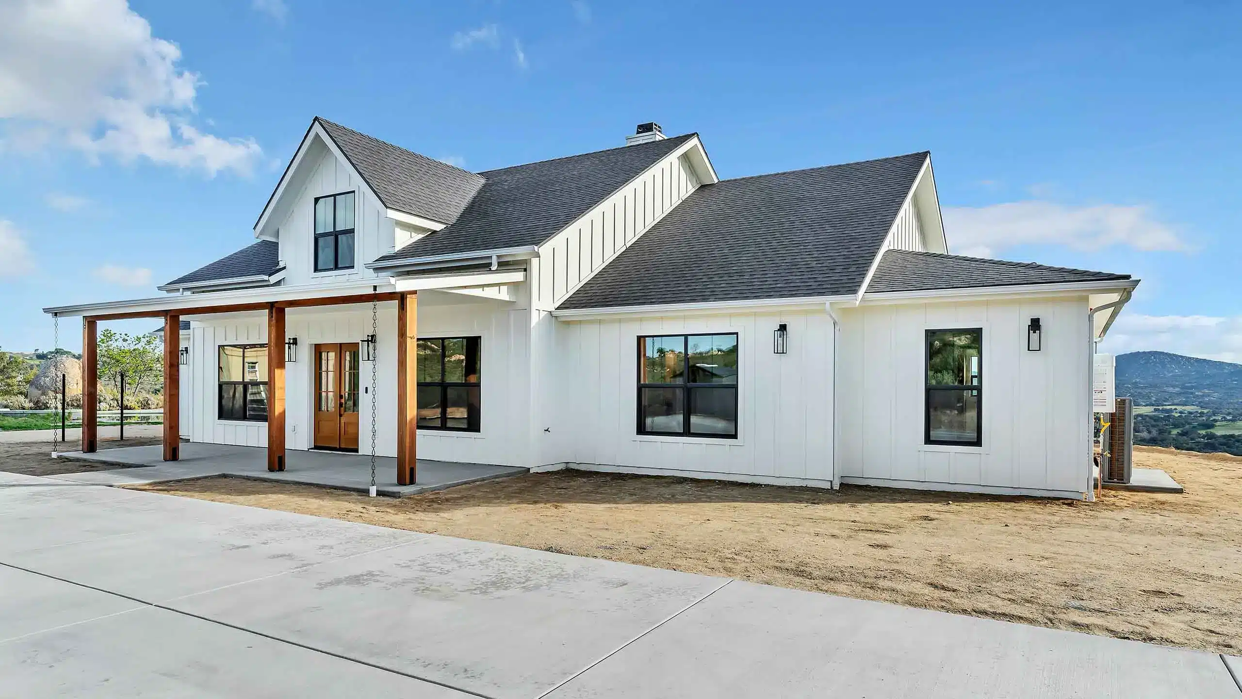 White modern farmhouse with gray gable roof, large windows, and a wooden-pillar porch under a blue sky.