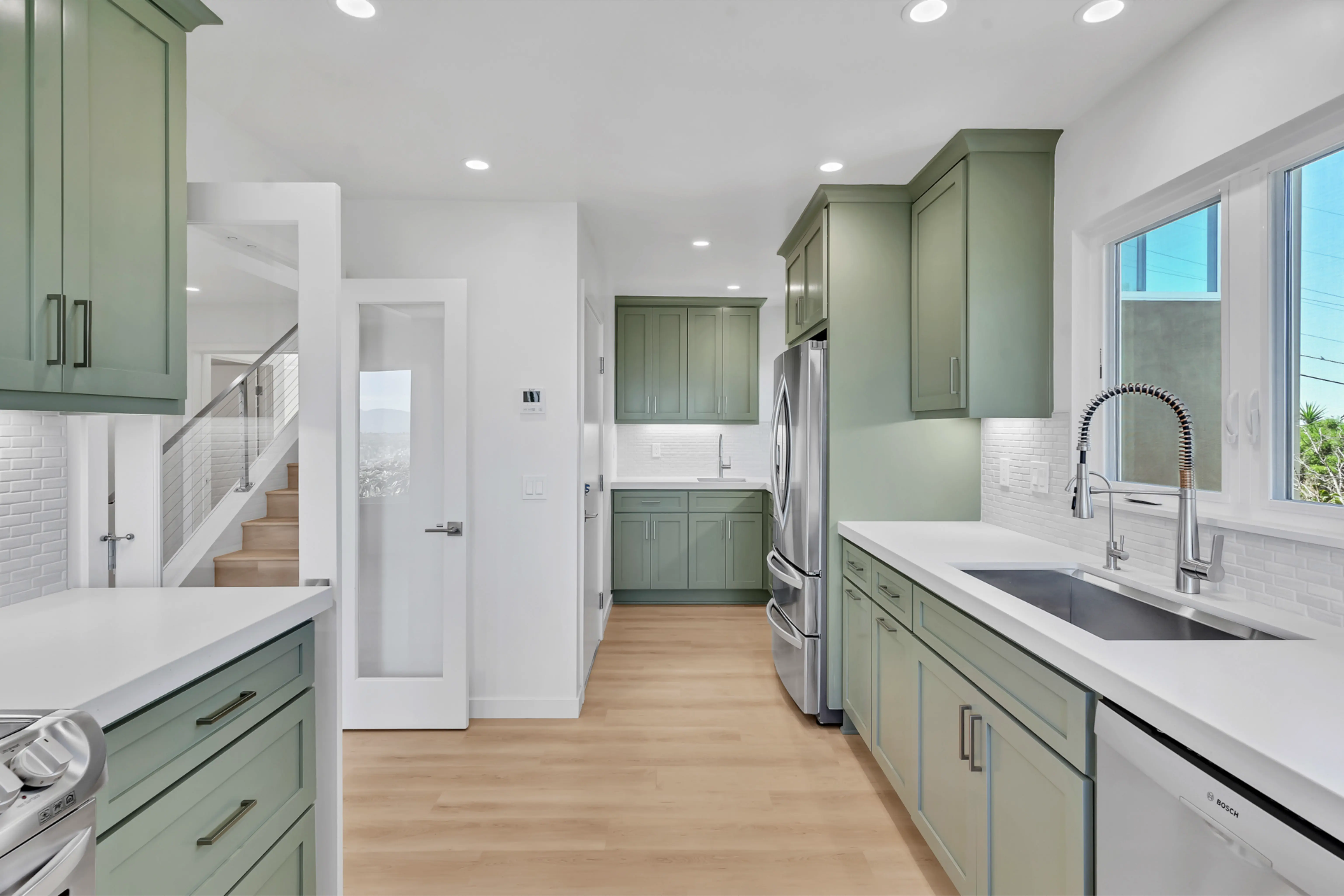 Bright modern kitchen with pale green cabinets, white countertops, and stainless steel appliances along a windowed wall.