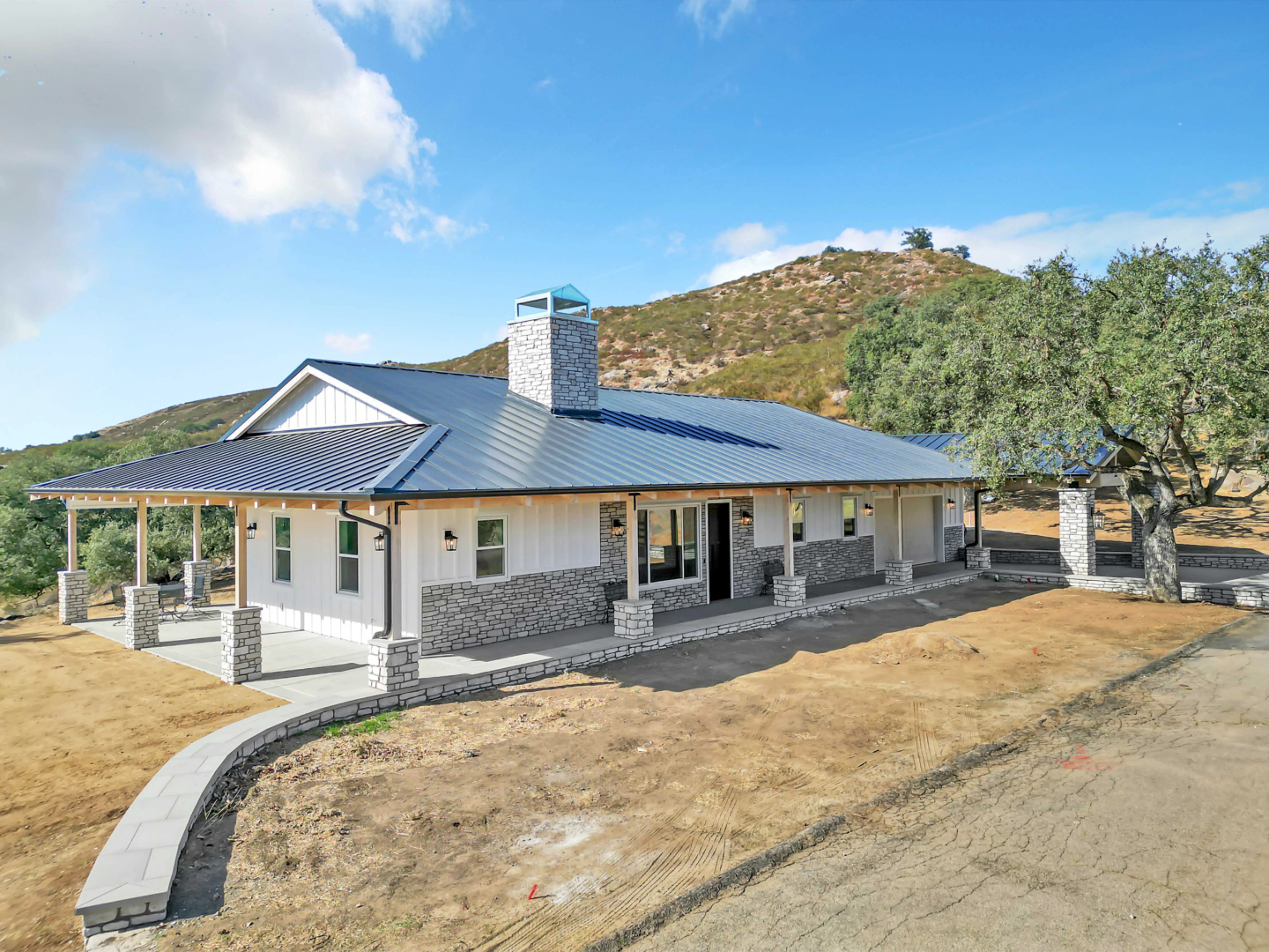 Modern single-story house with stone accents and a metal roof, chimney, and surrounding dirt yard against a hill and blue sky.
