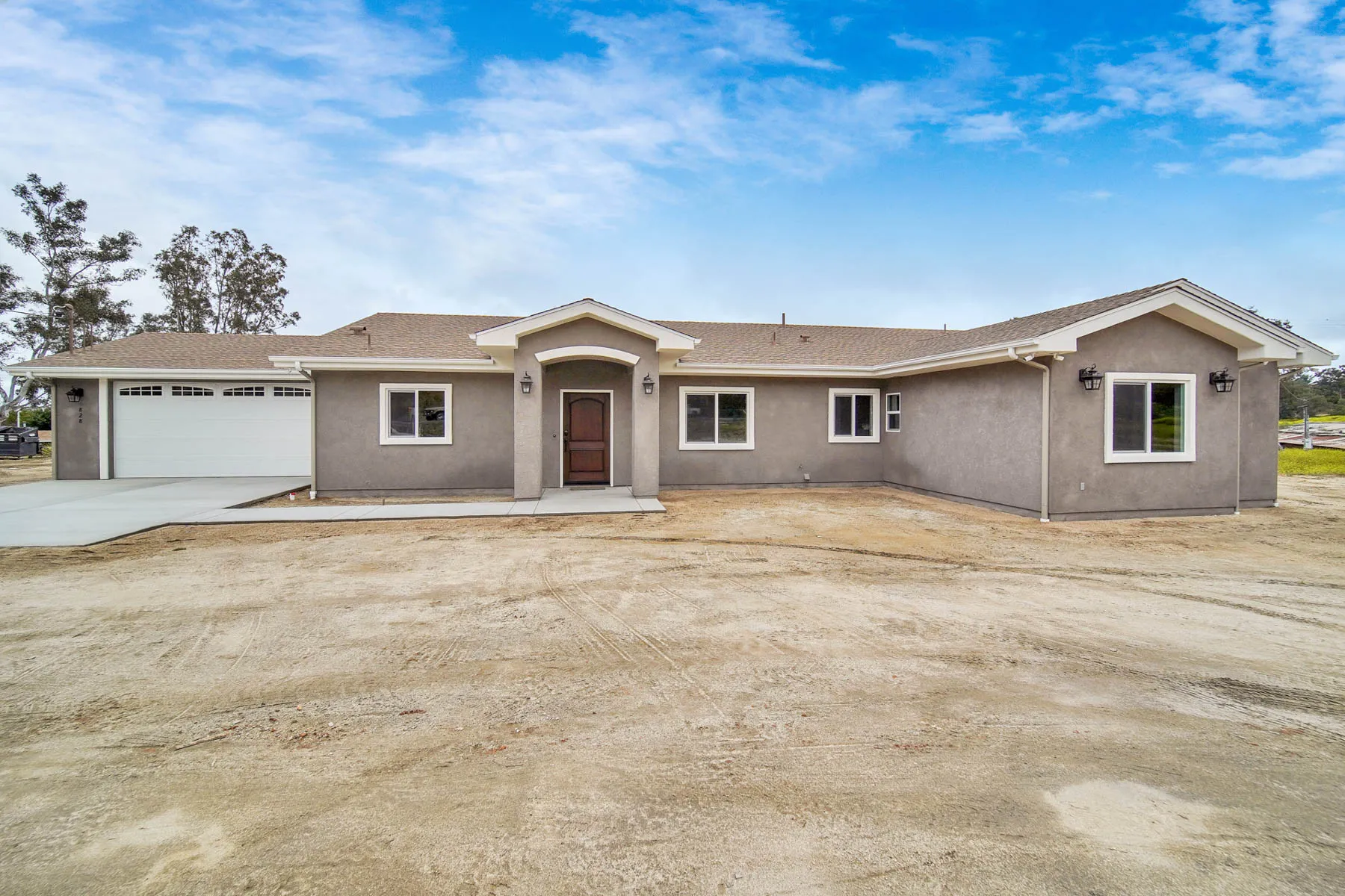Single-story gray stucco house under construction with a two-car garage and centered front door under an arched portico, dirt front yard and clear blue sky.