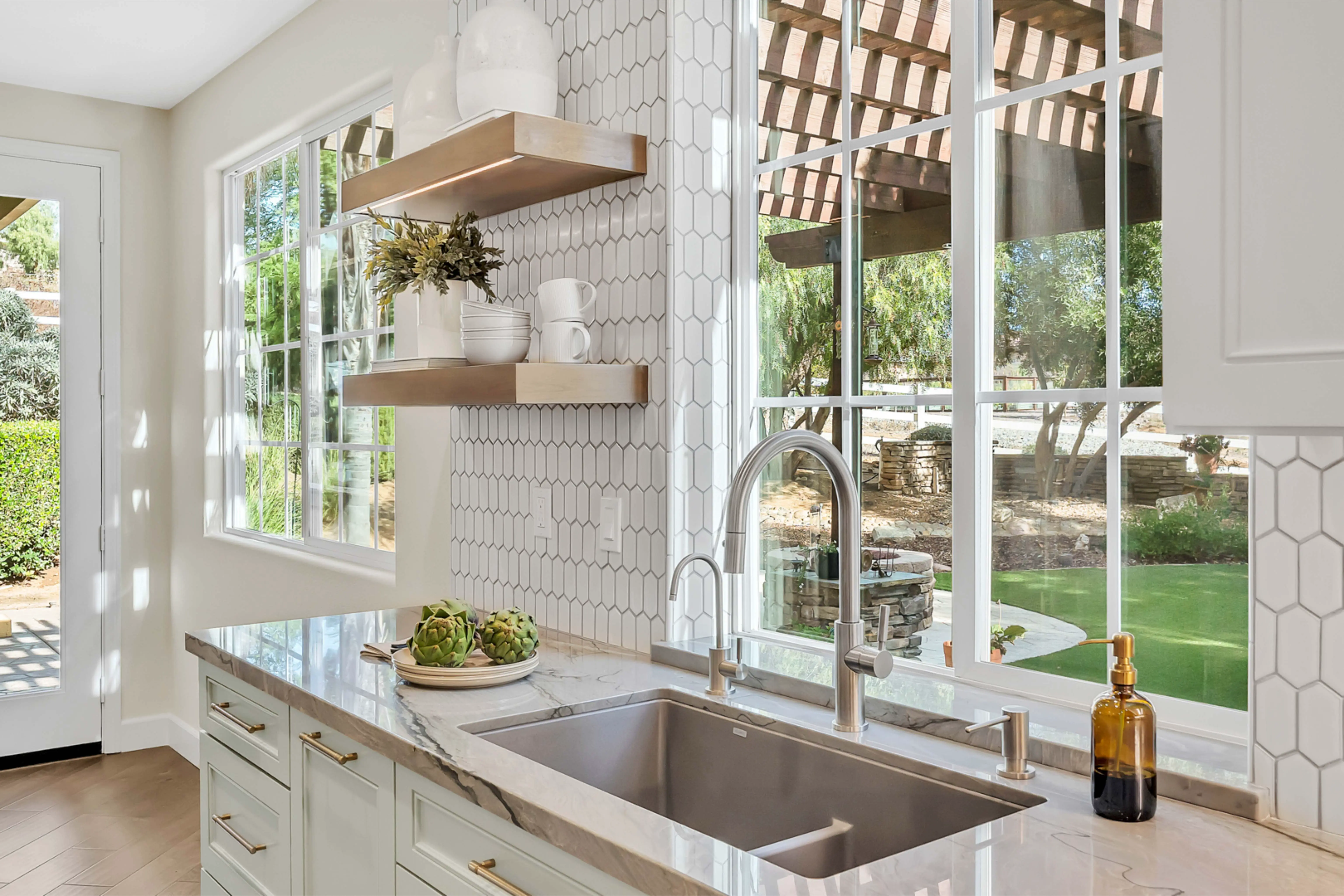 Bright kitchen with marble countertop, light cabinetry, hexagonal tile backsplash, and large windows overlooking a garden.