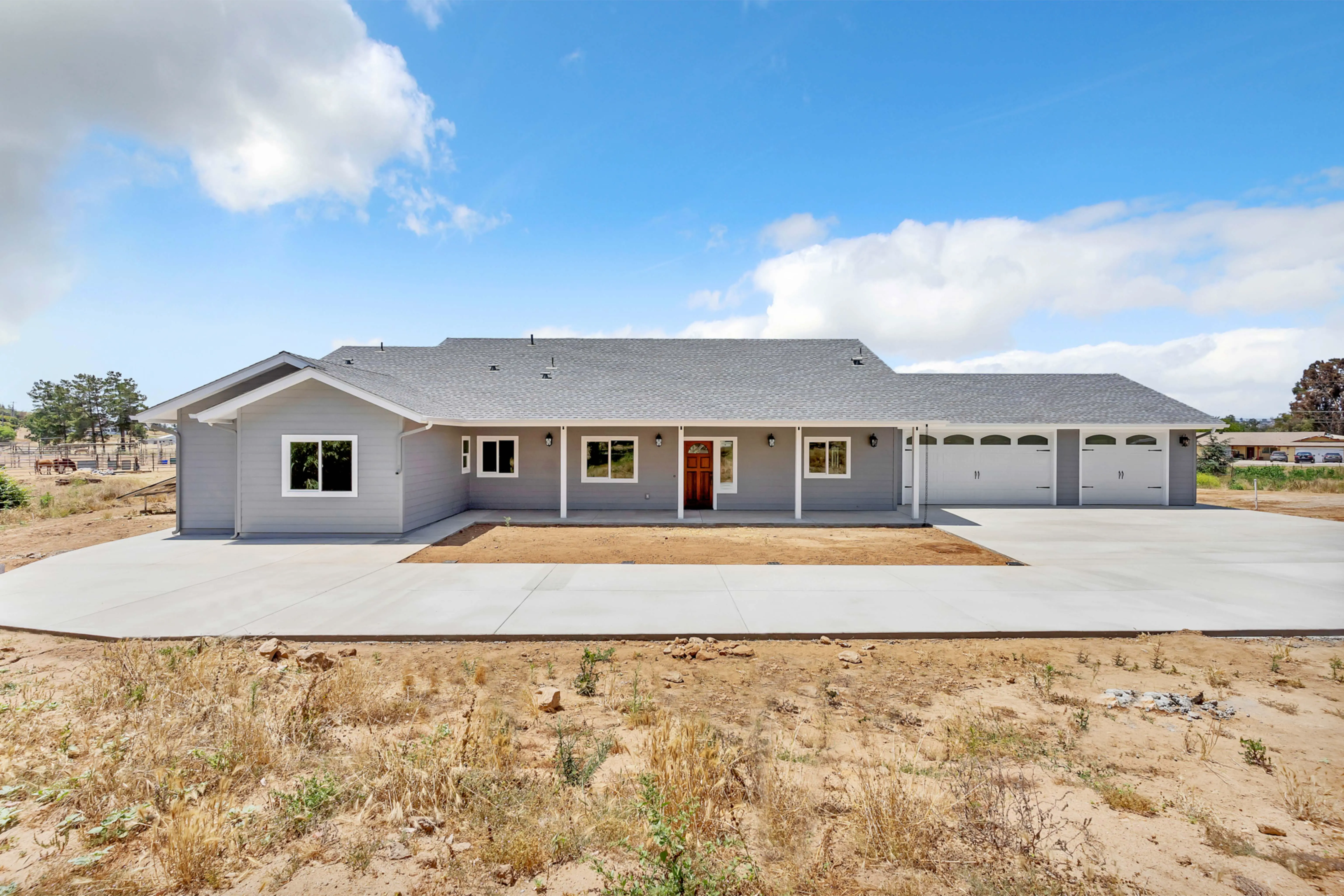 Front view of a single-story gray ranch-style house with a covered porch and a three-car garage, on a dirt lot under a blue sky.